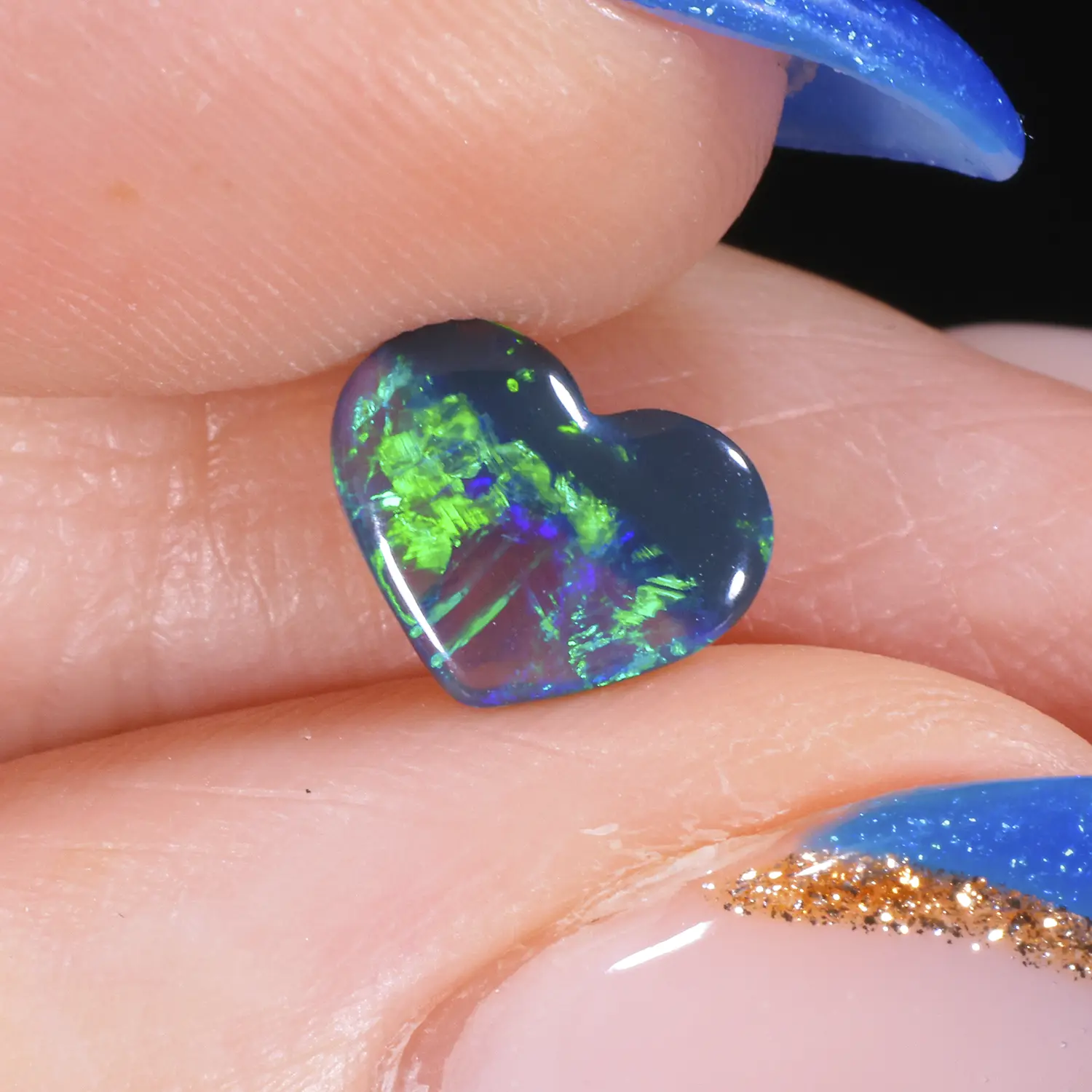 0.54ct untreated black opal heart held between fingers in close-up, showing bright electric green and violet colour flash with feather patterning against a dark body, Lightning Ridge Australia