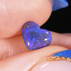 0.8ct solid black crystal opal heart held between fingers, displaying vivid blue and violet play-of-colour against a dark body tone, Lightning Ridge Australia