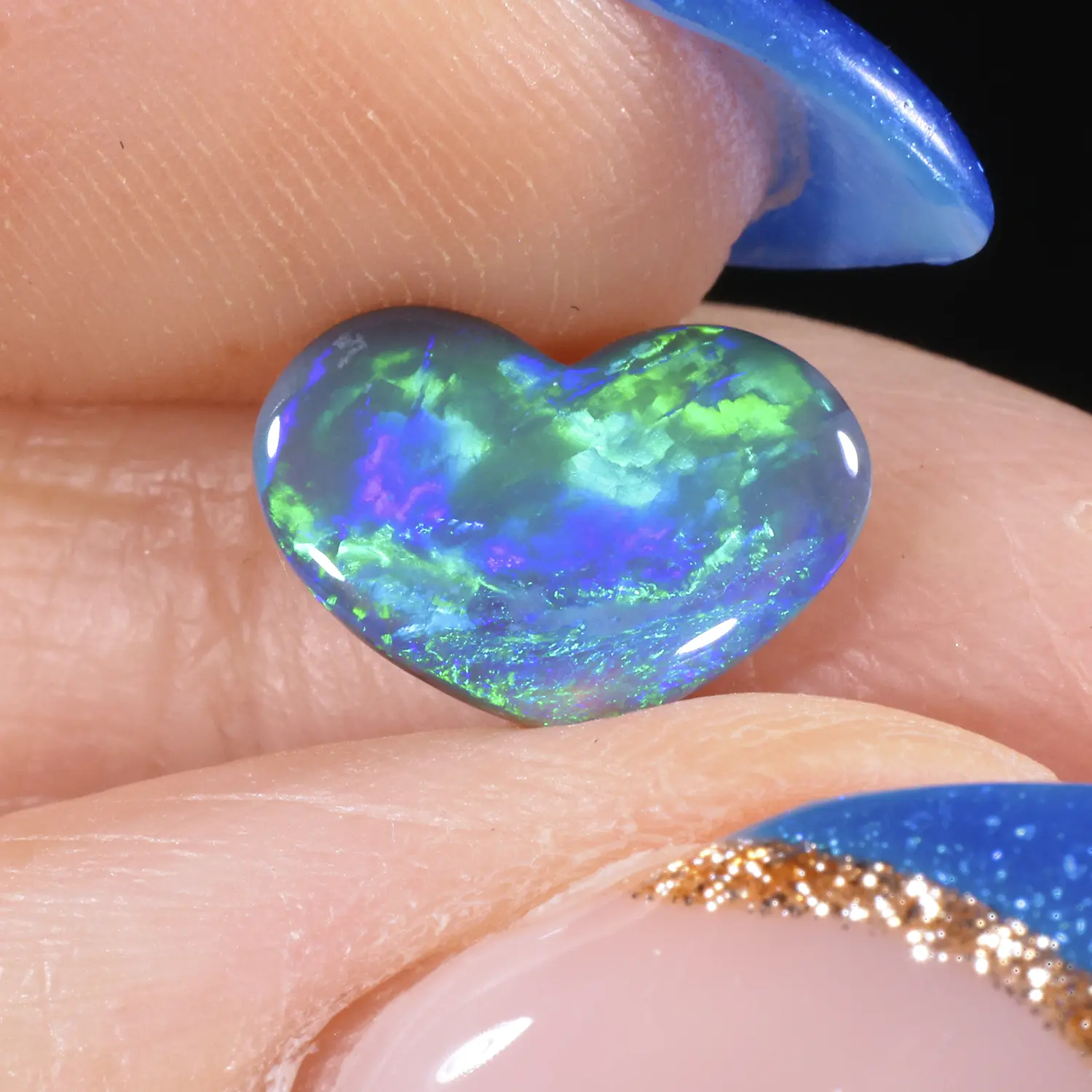 0.96ct solid black opal heart held between fingers in close-up, displaying vivid electric green and blue play-of-colour with mosaic patterning against a dark body tone, Lightning Ridge Australia