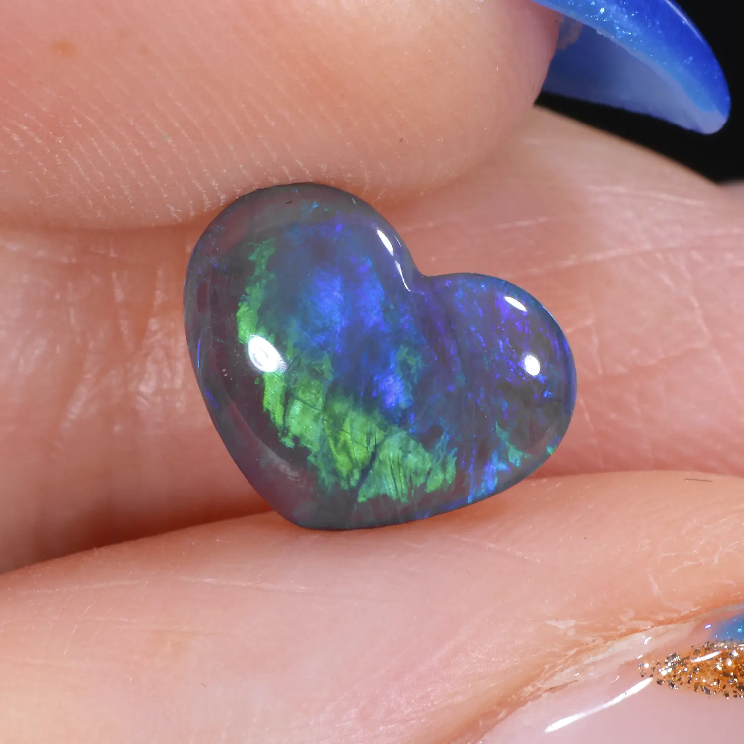1.11ct untreated black crystal opal heart held between fingers, showing vivid electric green and blue colour flash with a feather-like pattern against a dark body tone, Lightning Ridge Australia