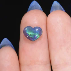 1.11ct natural black crystal opal heart resting on a fingertip, displaying vivid green and blue play-of-colour against a dark translucent body, Lightning Ridge Australia