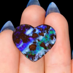 16.5ct untreated boulder opal heart shape resting across fingers, exhibiting bright blue, purple and electric green colour play with ironstone inclusions, Winton Queensland Australia